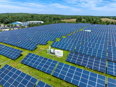 Aerial view of solar photovoltaic power plant on a capped landfill with rows of blue panels in a field, surrounded by trees under a bright sky. Massachusetts, USA.