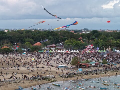 Aerial view of Mertasari Beach during the Bali Kite Festival with colorful kites flying over a crowded shore, traditional boats, and lush greenery in Denpasar Selatan, Bali, Indonesia.