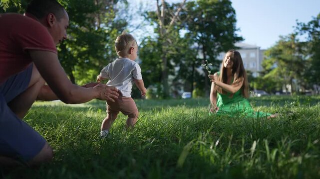 Father helps son walk toward mom in park. Happy son takes first steps with father and mom cheering in park. Family bonding as mom father guide son. Joyful outdoor moment with son mom father in park.