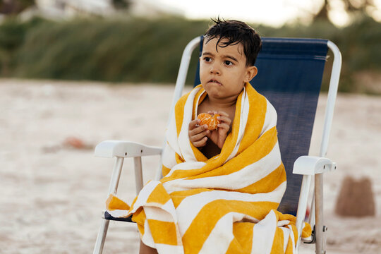 A young boy wrapped in a yellow striped beach towel sits on a folding chair at the seaside, holding a peeled orange and looking thoughtful after a swim.