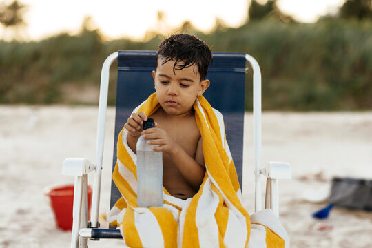 A thoughtful young boy sits wrapped in a yellow striped towel on a beach chair, holding a water bottle after swimming. Wet hair, sand toys and a calm seaside atmosphere convey summer relaxation.