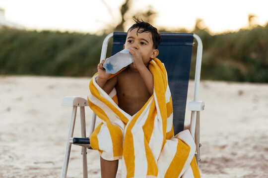 A young boy wrapped in a yellow-and-white striped towel sits on a beach chair, drinking from a water bottle with wet hair, relaxed and hydrated after play at the sandy seaside.