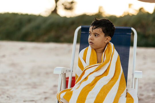 Pensive Young Boy Wrapped in Yellow Striped Towel Sitting on Beach Chair at Sunset
