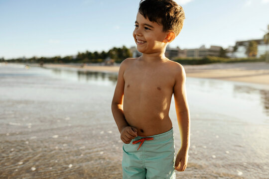 Smiling young boy in swim trunks stands on a sunlit beach shoreline, enjoying a carefree summer vacation at a tropical resort. Warm light, wet sand and gentle waves create a joyful, relaxed mood.