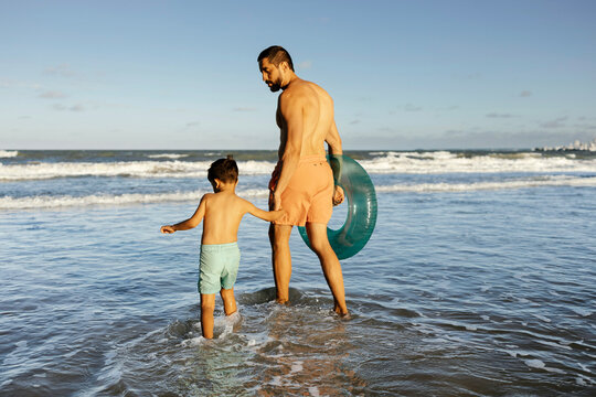 A father and young son walk hand in hand into gentle ocean waves, carrying an inflatable tube on a warm summer afternoon, capturing family bonding, relaxation and seaside fun.