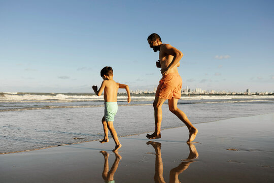 Father and son run and play along a sunny shoreline, barefoot on wet sand with reflections in shallow water. Warm light evokes joy, connection, health, and carefree summer family bonding at the beach.