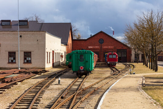 View of the Aukstaitija narrow gauge railway depot with vintage green carriages and a red locomotive on tracks beside historic brick buildings in Panevezys, Panevezio apskritis, Lithuania.