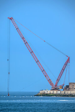 View of a heavy-duty lattice-boom crawler crane positioned on a concrete breakwater extending into the sea under a clear blue sky in Matosinhos, Porto District, Portugal.