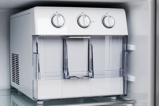 Portable ice maker sits inside clean refrigerator compartment. Unit makes ice cubes for cold drinks. Transparent bin holds frozen water. Modern kitchen appliance.