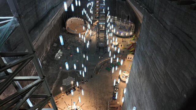 Tilt-down shot revealing the massive depth of the Salina Turda salt mine cavern in Cluj County, Romania, showing the illuminated underground lake, recreational wooden structures, and dark geology
