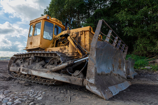 View of a yellow bulldozer parked on a dirt field with trees in the background under a cloudy sky in Kelme, Siauliai County, Lithuania.
