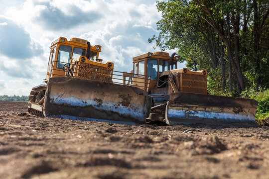 View of two yellow bulldozers parked on a dirt field with trees and a cloudy sky in the background in Kelme, Siauliai County, Lithuania.