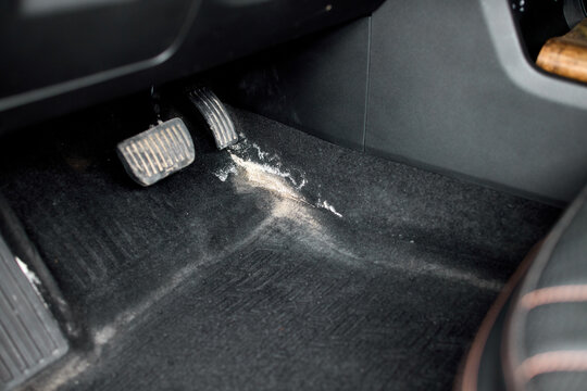 Close view of vehicle driver footwell showing scattered white powder on dark carpet near brake and accelerator pedals under soft interior light, with car, vehicle, interior, footwell detail