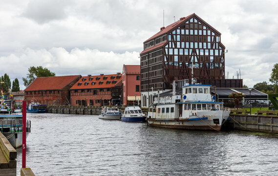 View of the Dane River waterfront featuring historic red-brick buildings, a modern glass-fronted structure, and moored boats under a cloudy sky in Klaipeda, Klaipeda County, Lithuania.