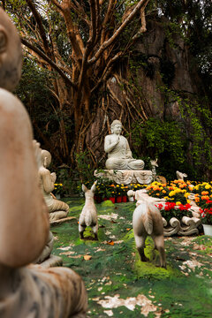 View of The Marble Mountains featuring a serene Buddha statue surrounded by stone animal figures, vibrant marigolds, and lush trees set against a rocky cliff in Da Nang, Da Nang, Vietnam.