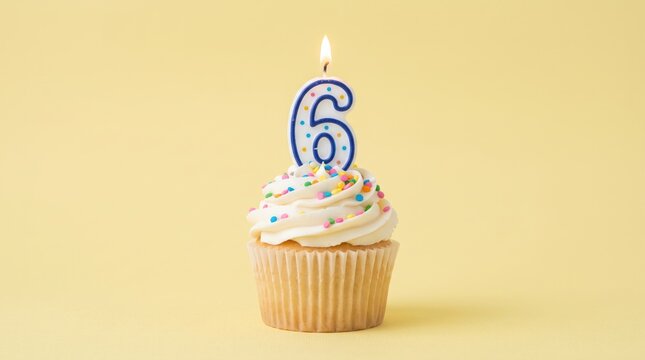 Birthday cupcake with white frosting and sprinkles topped by a lit number six candle, isolated on a soft yellow backdrop