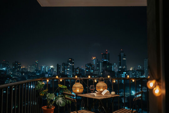 A cozy balcony at night with string lights a table two chairs potted plants lanterns a glass of red wine an open book and a smoking incense stick overlooking a vibrant city skyline