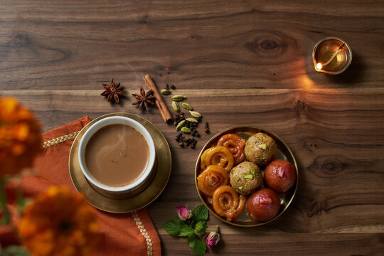A steaming cup of Indian chai tea on a brass saucer with a plate of assorted Indian sweets jalebi ladoo gulab jamun aromatic spices a lit diya oil lamp and rosebuds