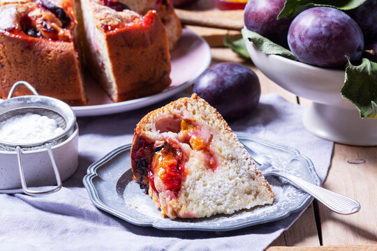 Plum pie with filling and sugar crust, sprinkled with powdered sugar and plums on a wooden table.