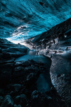 View of a deep blue ice cave with smooth glacial walls and dark volcanic rocks surrounding a stream of water in Iceland.