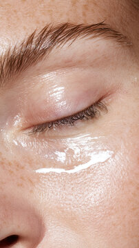 Person's closed eye and surrounding skin showing visible freckles. Featuring a clear. Glossy gel or serum applied to the eyelid. Emphasizing a radiant. Hydrated