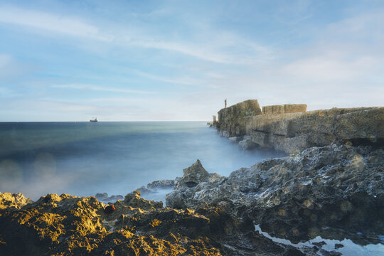 View of a person standing on a stone breakwater extending into the sea with rocky foreground and a distant ship under a blue sky in San Pedro de Macoris, San Pedro de Macoris, Dominican Republic.