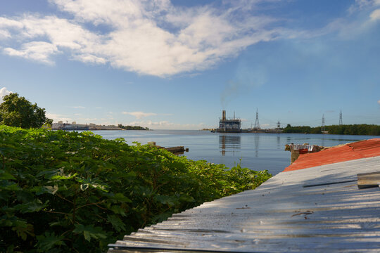 View of an industrial facility with smoke rising over coastal waters, framed by a corrugated metal roof and lush foliage in San Pedro de Macoris, San Pedro de Macoris, Dominican Republic.