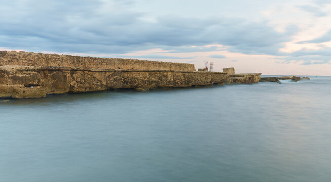 View of the stone seawall along the coast with calm water and a cloudy sky at dusk in San Pedro de Macoris, San Pedro de Macoris, Dominican Republic.