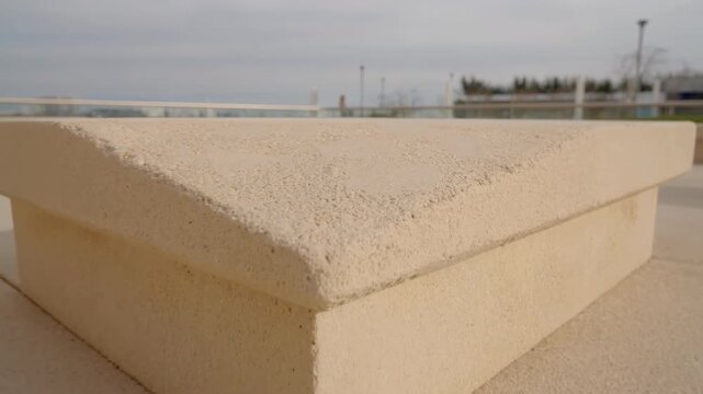 Low-angle close-up view revealing beige concrete capstone on outdoor plaza plinth, with chamfered top, textured paving, soft shadows, and minimal modern architectural detail