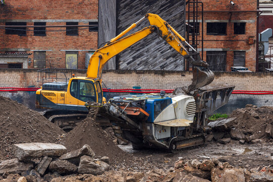 View of a yellow excavator and a mobile rock crusher working at a construction site with piles of dirt and a brick building in Vilnius, Vilnius County, Lithuania.