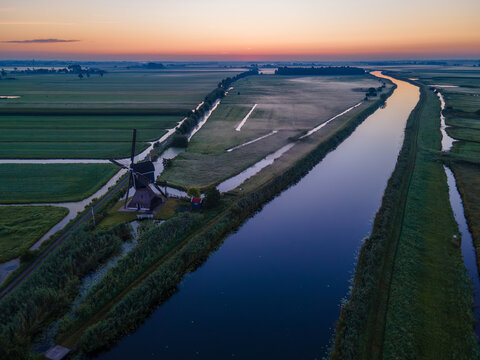 View of a traditional Dutch windmill standing beside a calm canal in the countryside at sunrise, with lush green fields and soft morning light in the Netherlands.