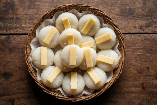 Top view of traditional Filipino white puto rice cakes topped with cheese slices inside woven basket. Steamed sweet treats arranged on wooden surface for dessert snack. Authentic homemade southeast
