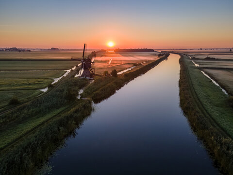 View of a traditional Dutch windmill standing beside a calm canal during a golden sunrise, with expansive green fields and misty meadows in the Netherlands.