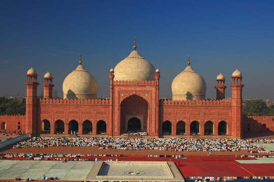 View of the historic Badshahi Mosque with its iconic red sandstone facade and white domes, featuring a large courtyard filled with people praying on red carpets in Lahore, Punjab, Pakistan.