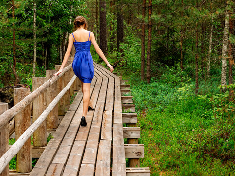 Woman in blue dress walks on wooden forest bridge. She smiles and poses near railing. Green pine trees surround the path. Scene shows natural daylight in summer. Young slim model. Prom photo.