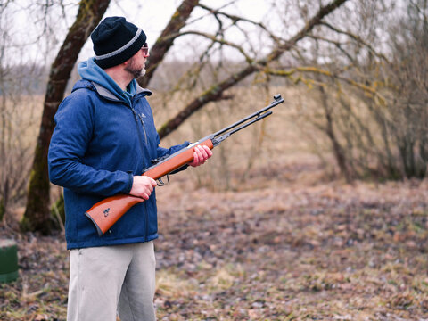 Person aims rifle outdoors in wooded area. Bare branches frame the scene. Ground covered with fallen leaves. Season appears late fall or early winter. No animals or plants identified