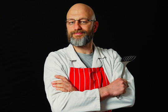 Bald man with glasses and beard stands confidently. He wears white coat and red striped apron. Holds spatula in hand. Smiles at camera. Black background highlights his figure