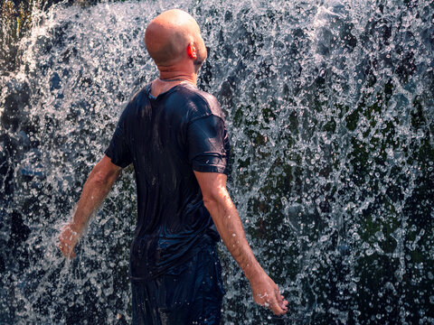 Man stands barefoot in shallow water before a cascading waterfall. Sunlight shines behind him, highlighting falling water droplets. He wears casual clothing, smiling toward camera. Summer heat concept