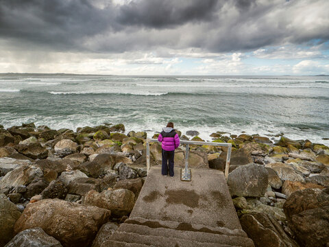 Child in a purple jacket stands on stone steps by a rocky shoreline. Waves crash against large, wet boulders under a cloudy sky. The child looks out at the ocean, Strandhill, Sligo, Ireland. Travel
