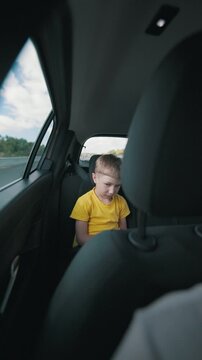 Young boy in yellow shirt sits quietly in the back seat of a car, visible headrest and interior elements, with blurred scenery through the window