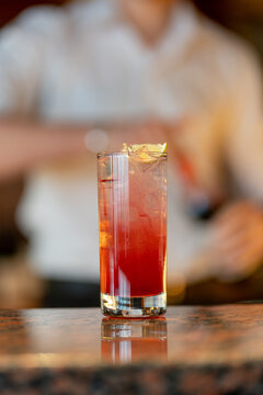View of a refreshing red cocktail in a tall glass with ice and a lime wedge on a wooden bar counter with a bartender in the background in Asker, Akershus, Norway.