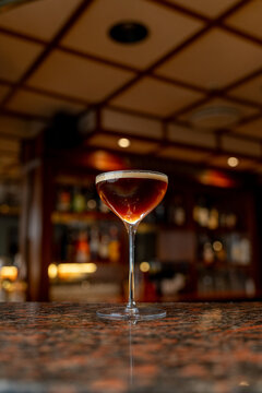 View of an espresso martini cocktail served in a stemmed glass on a polished granite bar counter with a blurred background of a dimly lit bar in Asker, Akershus, Norway.