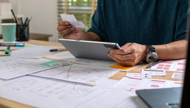UX UI designer working on wireframes for a website and mobile app prototype, surrounded by sketches of user-flow and design tools, in concept of website and mobile application design concept.