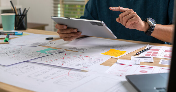 UX UI designer working on wireframes for a website and mobile app prototype, surrounded by sketches of user-flow and design tools, in concept of website and mobile application design concept.