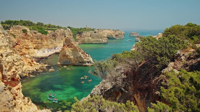 Kayakers explore turquoise waters surrounded by golden limestone cliffs, sea arches and a secluded beach on the Algarve coast of Portugal.