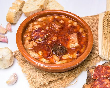 Traditional Spanish fabada stew served in a clay bowl with white beans, chorizo, morcilla, bacon, garlic cloves, sliced bread, and wheat ears