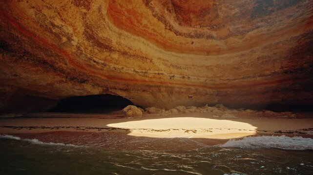 Sunlight illuminates a secluded sandy beach and layered limestone walls inside the famous Benagil Cave in Algarve, Portugal.