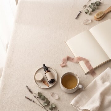 A serene still life arrangement features a dropper bottle with a blank label, a small dish, a wooden spoon, a cup of herbal tea, an open notebook, a ribbon, lavender sprigs, eucalyptus leaves.