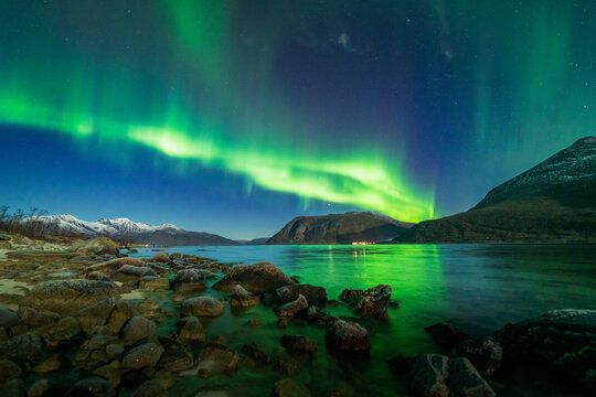 View of the northern lights illuminating the arctic fjord with rocky shoreline and snow-capped mountains in the background in Tromso, Troms, Norway.