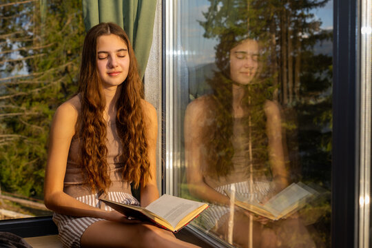 Young woman reading by sunlit window with reflection. Young woman sits by a sunlit window with an open book, eyes closed, while her reflection appears in the glass.
